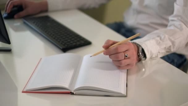 Επιχείρηση άνθρωπος που εργάζεται σε έναν υπολογιστή, καθιστώντας σημειώσεις σε ένα σημειωματάριο με ένα κοντινό πλάνο του στυλό — Αρχείο Βίντεο