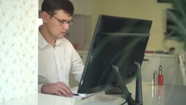 Um homem de camisa branca trabalha atrás de um computador em seu escritório em casa — Vídeo de Stock