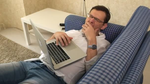 Der Mann mit Brille schlief während der Arbeit am Laptop auf seinem Sofa in seiner Stadtwohnung ein — Stockvideo