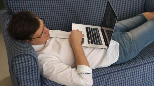 El hombre de gafas se quedó dormido mientras trabajaba en el portátil en su sofá en su apartamento de la ciudad — Vídeo de stock