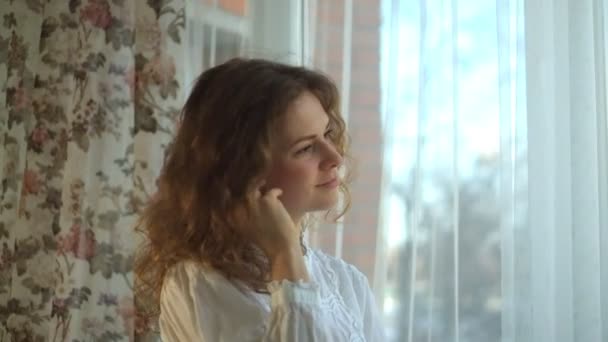 Кудрявые волосы девушка слушает музыку в наушниках у окна — стоковое видео