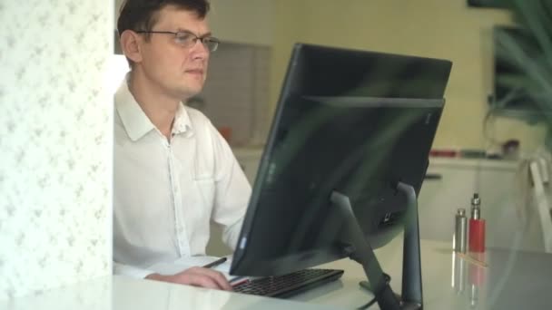 Um homem de camisa branca franze a testa, trabalha em um computador em seu escritório em casa — Vídeo de Stock