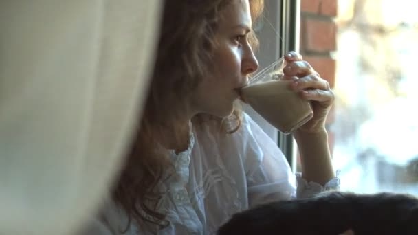 Menina em um cobertor quente, beber café com um gato no peitoril da janela . — Vídeo de Stock