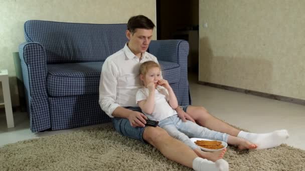 Hombre e hija viendo la televisión, sentados en el suelo comiendo bocadillos — Vídeo de stock