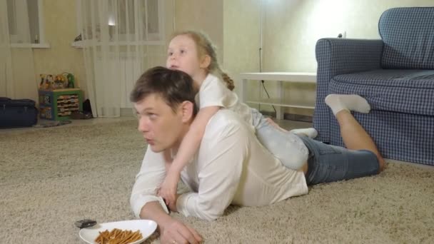 Hombre e hija viendo la televisión, sentados en el suelo comiendo bocadillos — Vídeos de Stock