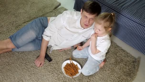 Hombre e hija viendo la televisión, sentados en el suelo comiendo bocadillos — Vídeo de stock