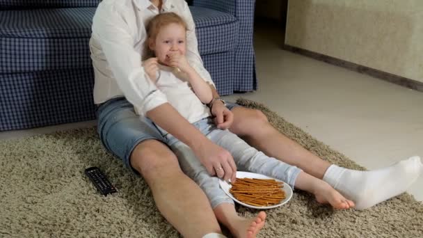 Homme et fille regardant la télévision, assis sur le sol manger des collations — Video