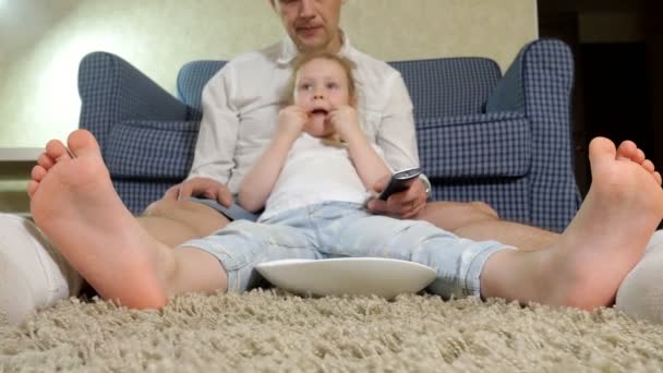 Мужчина и дочь смотрят телевизор, сидят на полу и едят закуски — стоковое видео