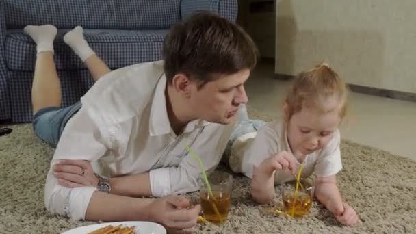 Мужчина и дочь смотрят телевизор, сидят на полу и пьют сок — стоковое видео