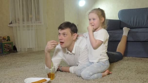 Homme et fille regardant la télévision, assis sur le sol manger des collations — Video