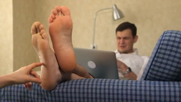 Ein ernsthafter Geschäftsmann liegt auf der Couch und arbeitet mit einem Laptop, jemand mit einer anderen Hand stört die Arbeit und berührt den Fuß. Füße, Nahaufnahme — Stockvideo
