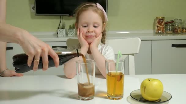 Egy aranyos kis lány ül egy asztalnál, úgy dönt, milyen ital, cola vagy gyümölcslé, az egészséges életmód fogalma — Stock videók
