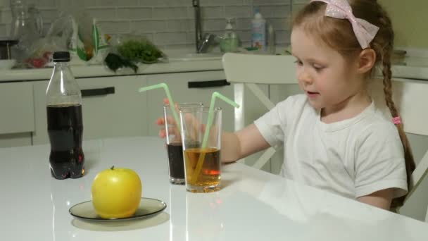 Una niña linda está sentada en una mesa, elige qué beber, cola o jugo, el concepto de un estilo de vida saludable — Vídeos de Stock