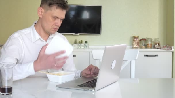 Um homem está sentado em uma mesa comendo macarrão chinês e trabalhando em um laptop — Vídeo de Stock