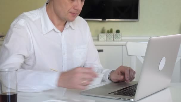 Un uomo è seduto a un tavolo a mangiare tagliatelle cinesi e lavorare su un computer portatile — Video Stock