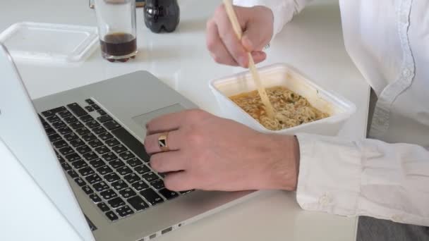 Um homem está sentado em uma mesa comendo macarrão chinês e trabalhando em um laptop — Vídeo de Stock