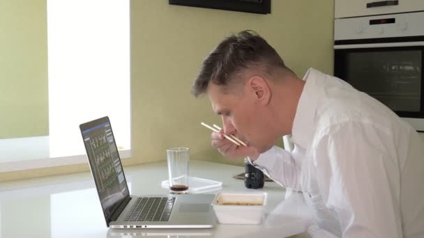 Egy férfi ül egy asztalnál eszik kínai tészta, és működő-ra egy laptop — Stock videók