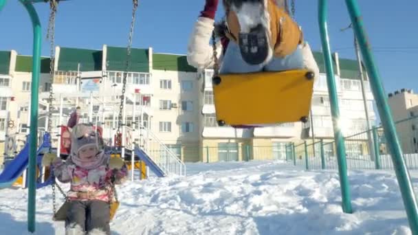 Jovem mãe com a criança balançando no balanço definido ao ar livre no parque de inverno. Neve caindo, nevasca, tempo de inverno — Vídeo de Stock