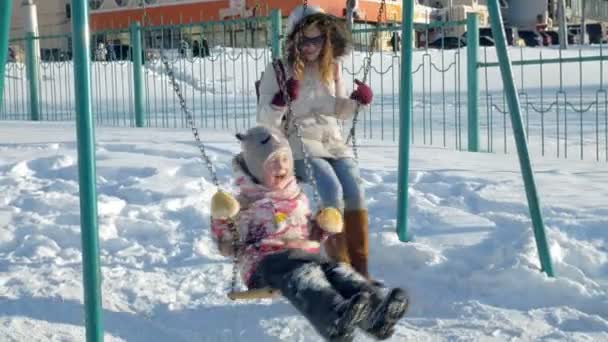 Молодая мама с ребенком качается на качелях на открытом воздухе в зимнем парке. Снегопад, снегопад, зимнее время — стоковое видео