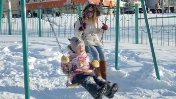 Ung mamma med barn som gungar på swing in utomhus i Vinterparken. Snö faller, snöfall, vintertid — Stockvideo