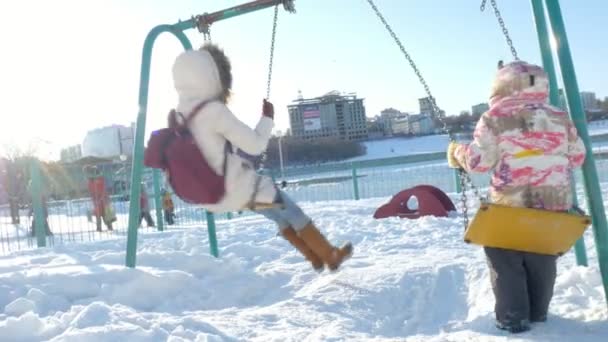 Νεαρή μητέρα με παιδί που αιωρούνται στην κούνια που υπαίθρια το χειμώνα πάρκο. Χιόνι που υπάγονται, χιονόπτωση, Χειμερινή ώρα — Αρχείο Βίντεο