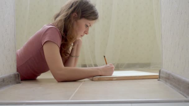 Giovane donna sdraiata sul pavimento e disegna una matita sulla tavoletta — Video Stock