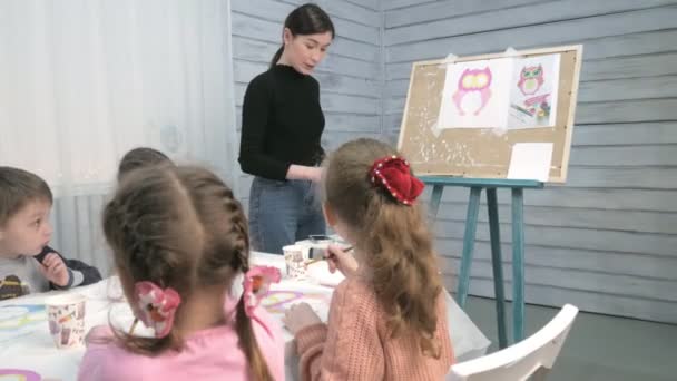 Bambini ragazzi e ragazze seduti insieme intorno al tavolo in classe e disegno. Con loro c'è la loro giovane e bella maestra . — Video Stock
