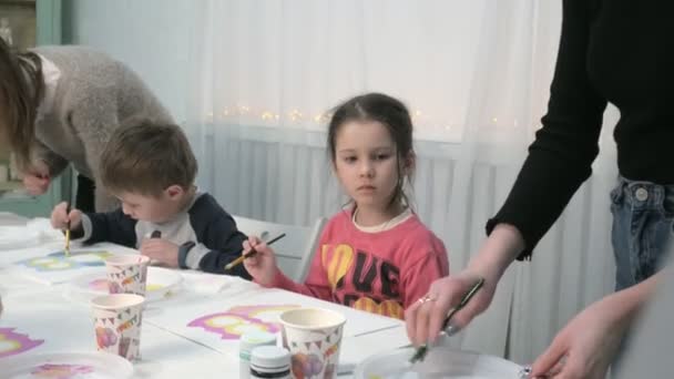 Niños niños y niñas sentados juntos alrededor de la mesa en el aula y dibujando. Con ellos está su joven y hermosa maestra . — Vídeo de stock
