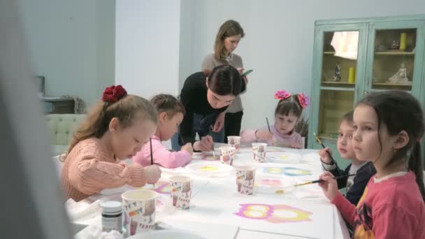 Дети сидят вместе за столом в классе и рисуют. С ними их молодой и красивый учитель . — стоковое видео