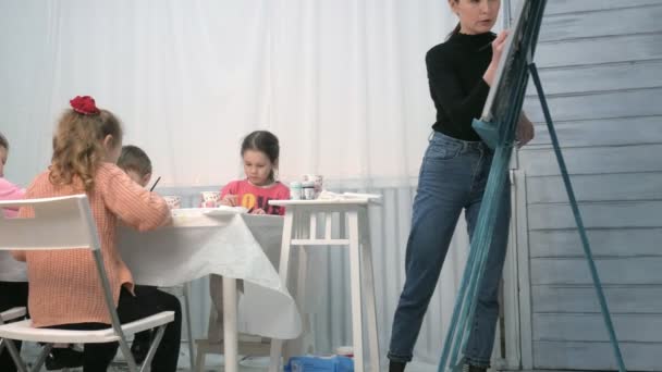 Kinderen-jongens en meisjes zitten samen rond de tafel in de klas en tekening. Met hen is hun jong en mooi leraar. — Stockvideo