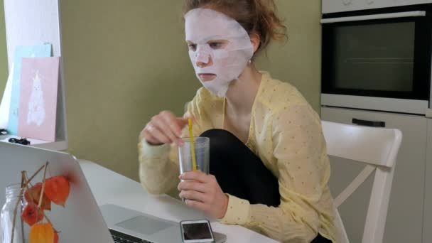 Jonge vrouw doet gezichtsmasker met zuivering masker, masker werken achter een laptop thuis — Stockvideo