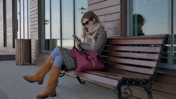 Mujer Usando Smartphone Relaja en el Banco en Beautiful Park. Mujer joven en la realización de gestos en la pantalla del teléfono. Tecnología al aire libre — Vídeos de Stock