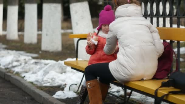 Uma menina bonita e sua mãe estão sentados em um banco de parque e comer uma deliciosa sobremesa e beber chá de uma garrafa térmica no início da primavera — Vídeo de Stock