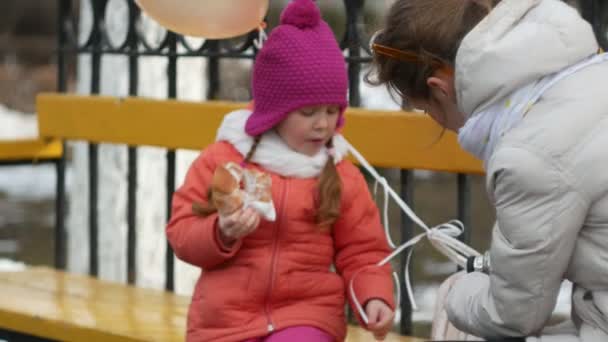 Una hermosa niña y su madre están sentadas en un banco del parque y comiendo un delicioso postre y bebiendo té de un termo a principios de primavera — Vídeos de Stock