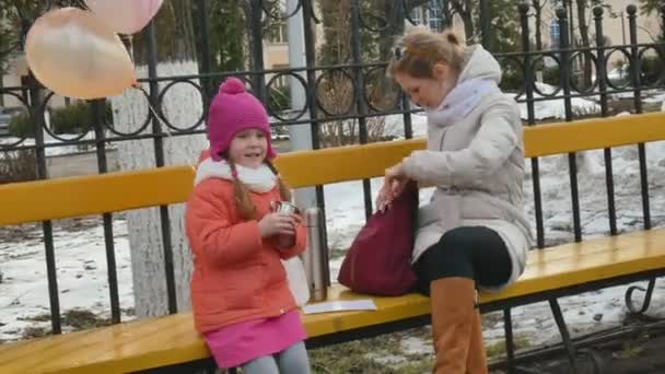 Una hermosa niña y su madre están sentadas en un banco del parque y comiendo un delicioso postre y bebiendo té de un termo a principios de primavera — Vídeo de stock