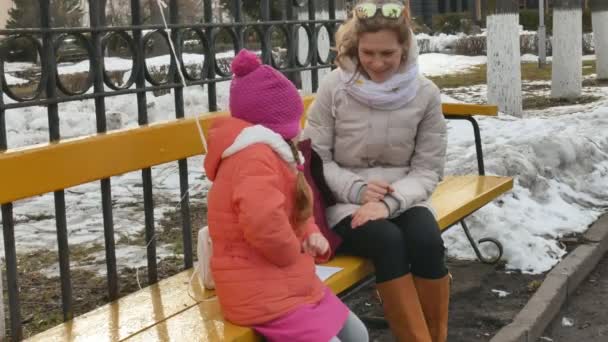 Una hermosa niña y su madre están sentadas en un banco del parque y comiendo un delicioso postre y bebiendo té de un termo a principios de primavera — Vídeos de Stock