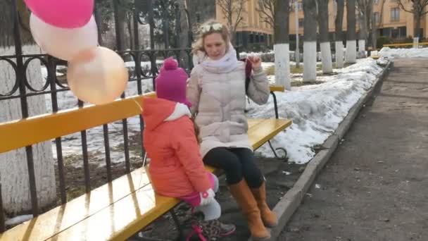 Una hermosa niña y su madre están sentadas en un banco del parque y comiendo un delicioso postre y bebiendo té de un termo a principios de primavera — Vídeos de Stock