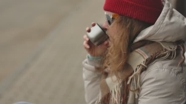 Joven hermosa mujer en un sombrero rojo con ropa de abrigo deportivo y rodillos, sentado en un banco de madera bebiendo té de un termo — Vídeos de Stock