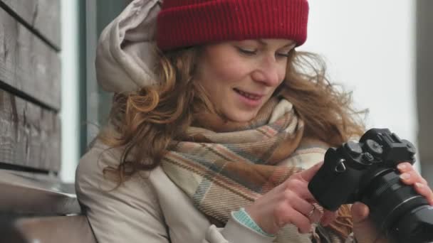 Jeune belle femme au chapeau rouge portant des vêtements chauds et sportifs et des rouleaux, assise sur un banc en bois et prenant des photos sur un appareil photo vintage — Video