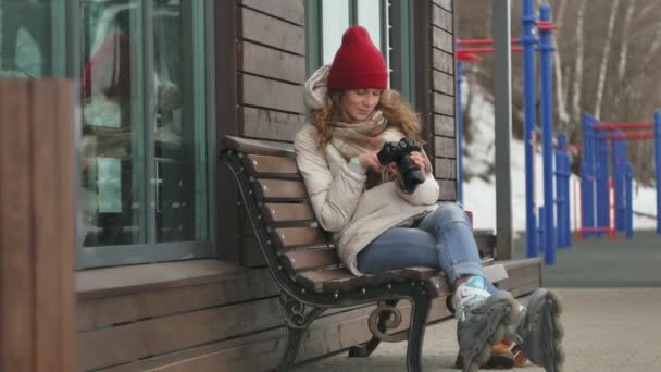 Giovane bella donna con un cappello rosso che indossa vestiti caldi sportivi e rulli, seduta su una panchina di legno e scattare foto su una fotocamera vintage — Video Stock