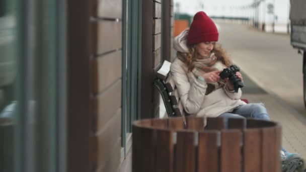 年轻漂亮的女人穿着红色的帽子身穿运动保暖衣服和滚筒, 坐在木凳上, 并在老式相机拍照 — 图库视频影像