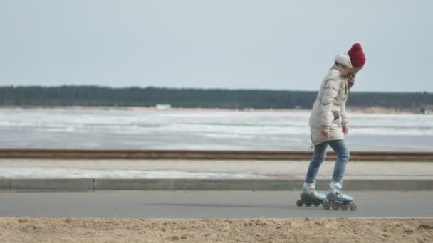 年轻漂亮的女人戴着红色的帽子, 穿着运动保暖的衣服和滚轮, 骑在海岸上的路上 — 图库视频影像