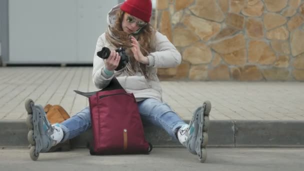Junge schöne Frau mit rotem Hut in sportlich warmer Kleidung und Rollschuhen, die auf der Asphaltstraße sitzt und mit einer Oldtimer-Kamera Fotos macht — Stockvideo