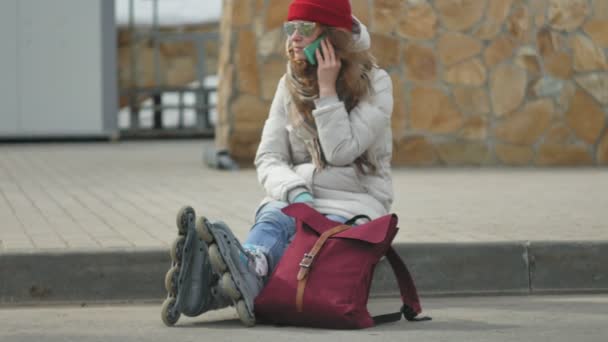 Молода красива жінка в червоній шапці носить спортивний теплий одяг і ролики, сидячи на асфальтовій дорозі і розмовляючи по телефону — стокове відео