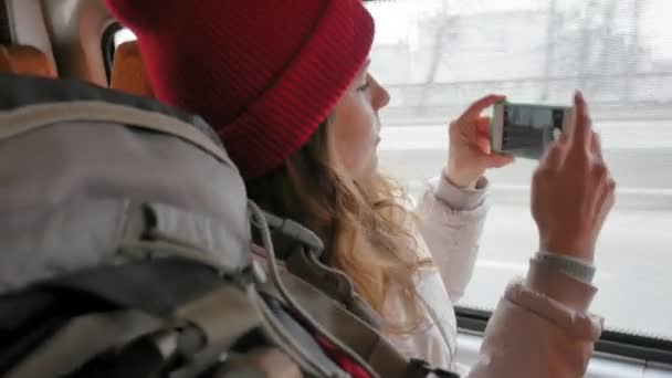 Joven mujer alegre en un sombrero rojo, viajando en autobús en un día triste. Ella toma fotos en un teléfono inteligente — Vídeos de Stock