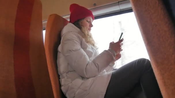 Μικρά χαρούμενα γυναίκα σε ένα κόκκινο καπέλο, ταξιδεύοντας με το λεωφορείο σε μια θλιβερή ημέρα. Παίρνει εικόνες σε ένα smartphone — Αρχείο Βίντεο