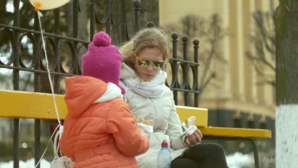 Una hermosa niña y su madre están sentadas en un banco del parque y comiendo un delicioso postre y bebiendo té de un termo a principios de primavera — Vídeo de stock