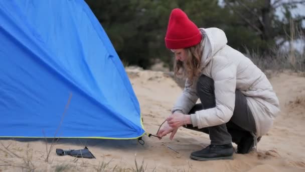 Attraente giovane donna turistica con un cappello rosso raccoglie una tenda turistica vicino alla foresta sulla costa — Video Stock