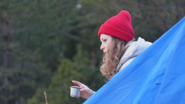 Atractiva turista joven en un sombrero rojo se sienta en una tienda de campaña turística y bebe té de un termo — Vídeos de Stock