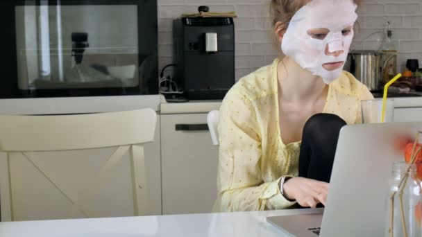 Jonge vrouw doet gezichtsmasker met zuivering masker, masker werken achter een laptop thuis — Stockvideo